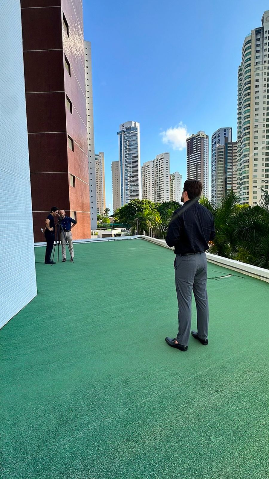 Bastidores de uma gravação no terraço: Walter de costas olhando os prédios, com a equipe e a câmera ao fundo