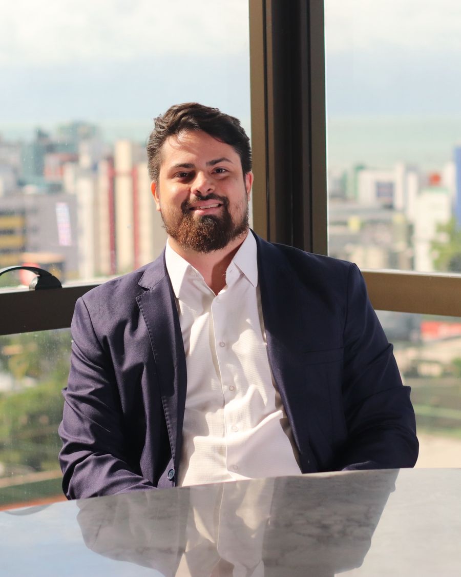 Walter Pimentel sorrindo, de blazer azul-marinho, sentado diante de uma janela com vista para a cidade